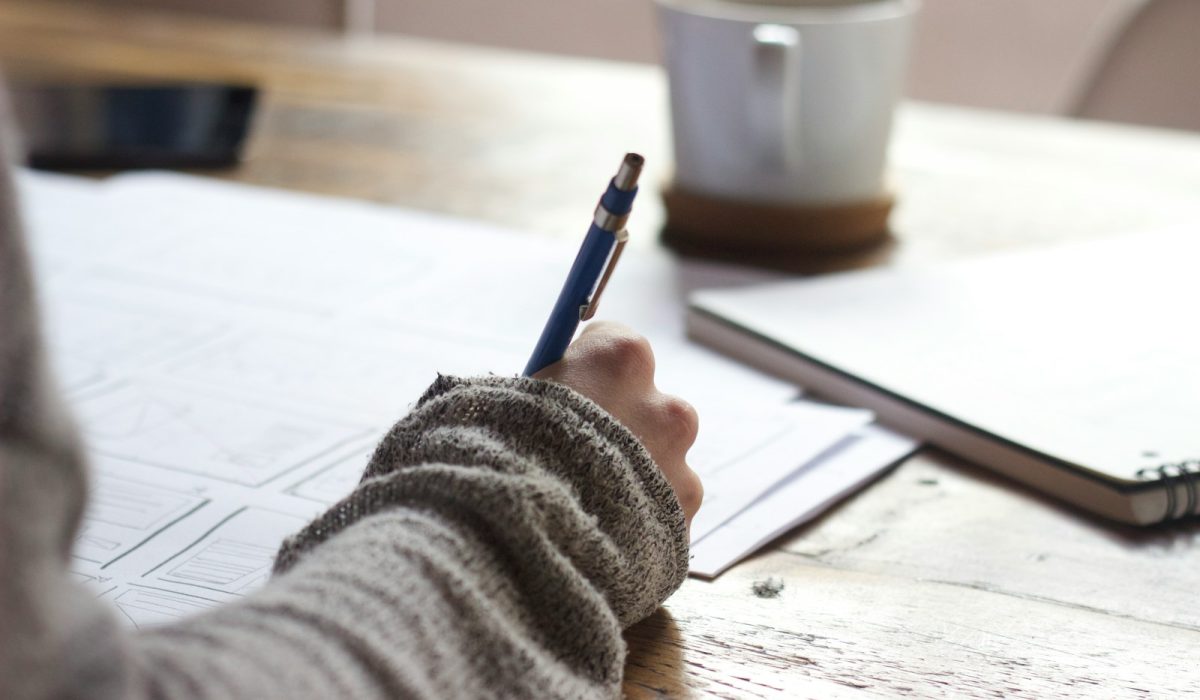 person writing on brown wooden table near white ceramic mug